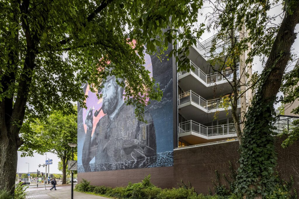 Muurschildering van een man met een pijp op een flatgebouw aan de Prins Hendrikkade, deels omgeven door bomen.